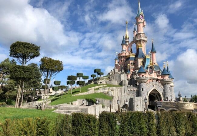 Zijkant van het Sleeping Beauty Castle in Disneyland Paris met groene heuvels, bomen en een blauwe lucht met wolken