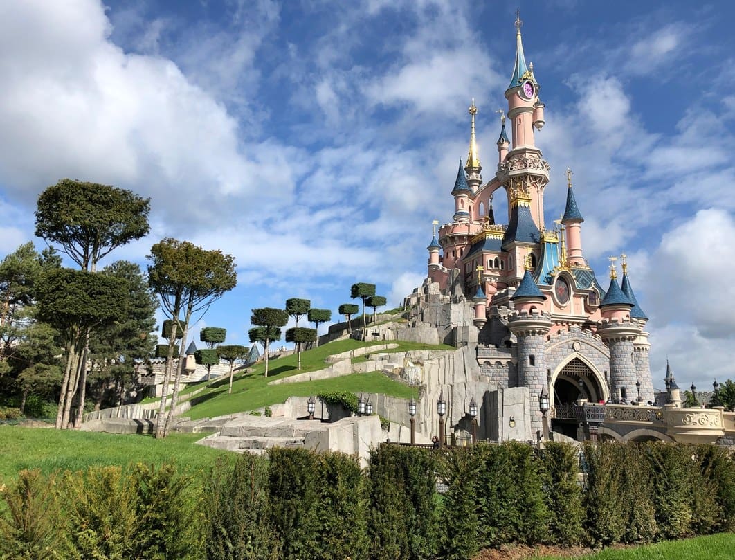 Zijkant van het Sleeping Beauty Castle in Disneyland Paris met groene heuvels, bomen en een blauwe lucht met wolken